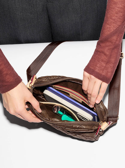 A person opens an MZ Wallace Small Metro Camera Bag in Walnut Oxford on a white surface, revealing a book, sunglasses, a pen, notebooks, cards, and small personal items neatly stored for convenient hands-free carry.