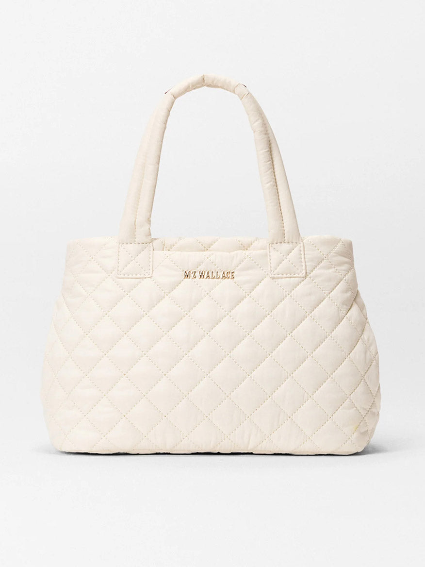 The MZ Wallace Small Metro City Tote in Sandshell Oxford is an off-white quilted handbag with two handles, Italian leather trim, and "MZ WALLACE" in gold letters on the front.