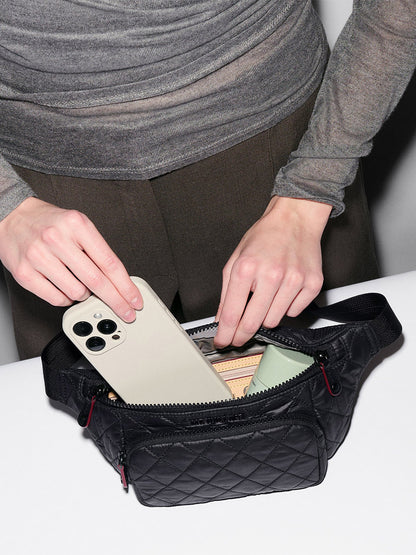 A person places a smartphone into an MZ Wallace Small Metro Sling in Black Oxford, which also holds a wallet and a green item.