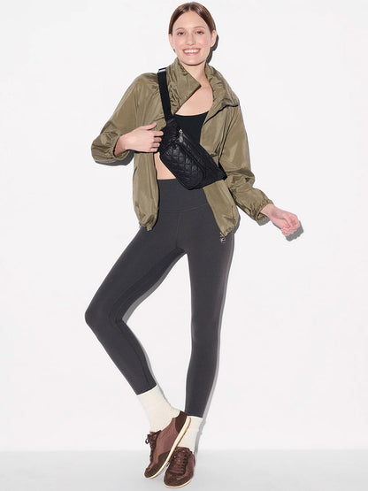 A person stands against a white background wearing a green jacket, black leggings, brown shoes, white socks, and the MZ Wallace Small Metro Sling in Black Oxford with Italian leather trim, smiling with one hand in a pocket.