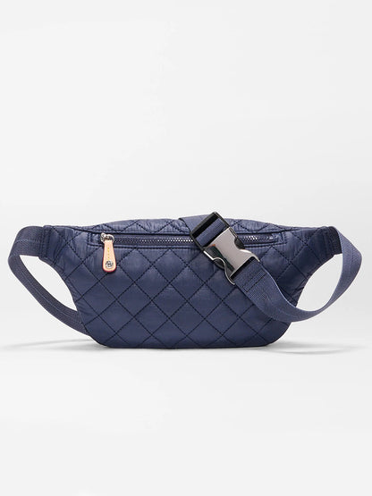 The MZ Wallace Small Metro Sling in Dawn Oxford is a navy blue quilted fanny pack with a front zipper pocket, adjustable crossbody strap, and plastic buckle, made from bluesign approved materials, shown on a plain white background.
