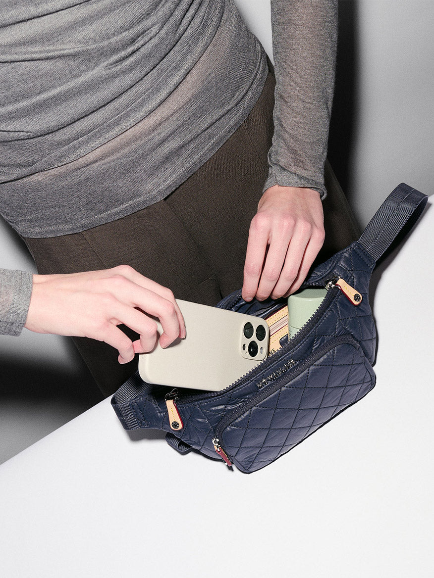 A person places a smartphone into the MZ Wallace Small Metro Sling in Dawn Oxford—a quilted navy waist bag with Italian leather trim—on a white surface, with a wallet and cards also visible inside.