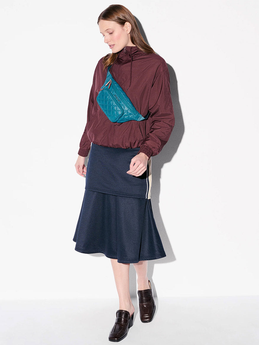 A woman stands against a white background wearing a maroon jacket, dark denim skirt, brown loafers, and carrying the MZ Wallace Small Metro Sling in Pacific Oxford with an adjustable crossbody strap.