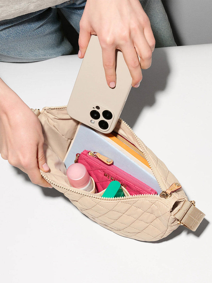 A person places a smartphone into the MZ Wallace Small Metro Smile Bag in Buff Oxford, which holds a pink zip pouch, a green item, a cylindrical container, and a notebook.