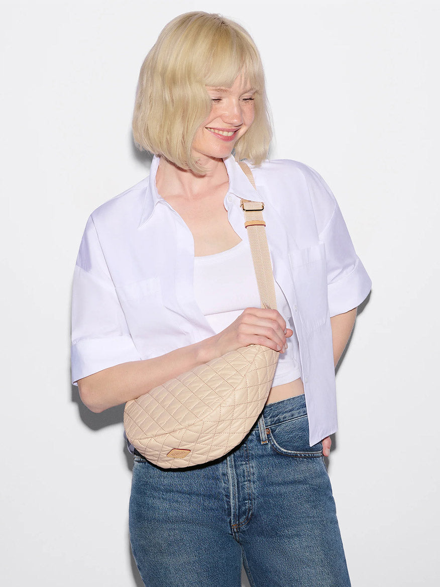A woman with short blonde hair in a white shirt, white tank top, and blue jeans holds the MZ Wallace Small Metro Smile Bag in Buff Oxford with an adjustable strap against a plain white background.