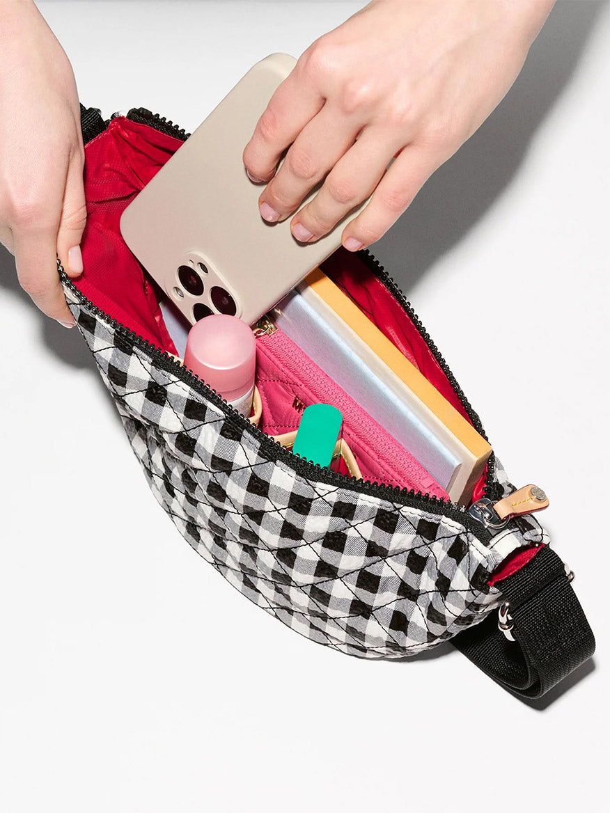 A hand places a smartphone into the MZ Wallace Small Metro Smile Bag in Black/White Gingham Oxford, featuring Italian leather trim, with a pink pouch, notebook, lip balm, and other small items inside.