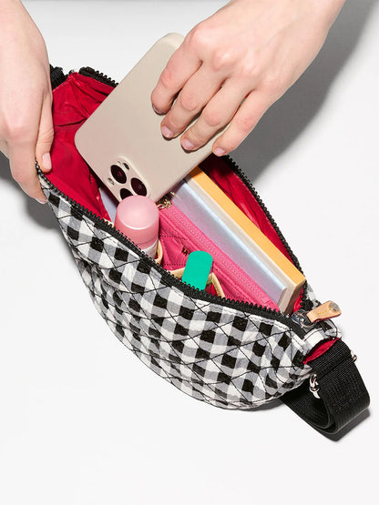 A hand places a smartphone into the MZ Wallace Small Metro Smile Bag in Black/White Gingham Oxford, featuring Italian leather trim, with a pink pouch, notebook, lip balm, and other small items inside.