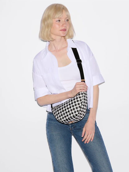 A woman in a white shirt and blue jeans stands against a plain backdrop, holding the MZ Wallace Small Metro Smile Bag in Black/White Gingham Oxford with an adjustable nylon strap.