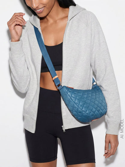 Woman wearing a gray zip-up hoodie, black sports bra, black biker shorts, and the MZ Wallace Small Metro Smile Bag in Atlantic Blue Oxford—crafted from recycled oxford nylon—standing against a plain white background; an ideal crossbody choice for everyday carry.