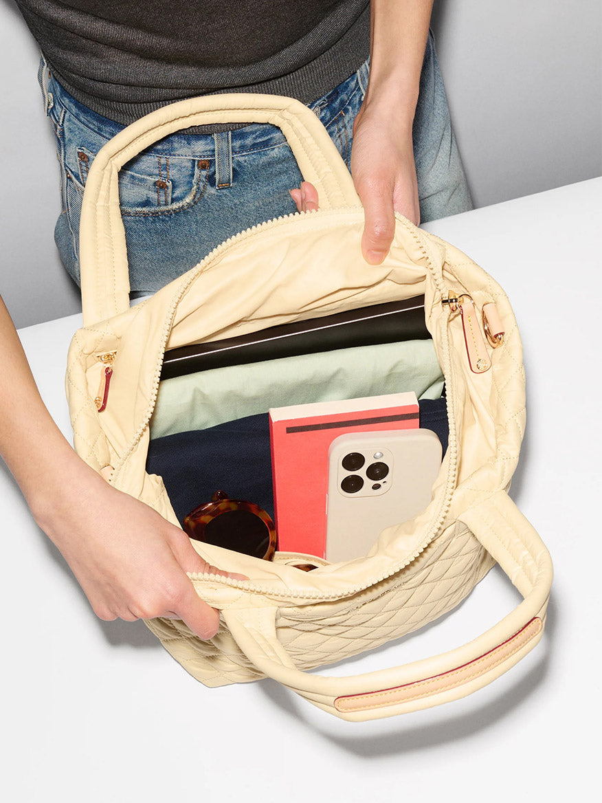 A person holds an open MZ Wallace Small Metro Tote Deluxe in Chamomile Oxford, showing a phone, sunglasses, notebooks, and a laptop inside its zip top closure.