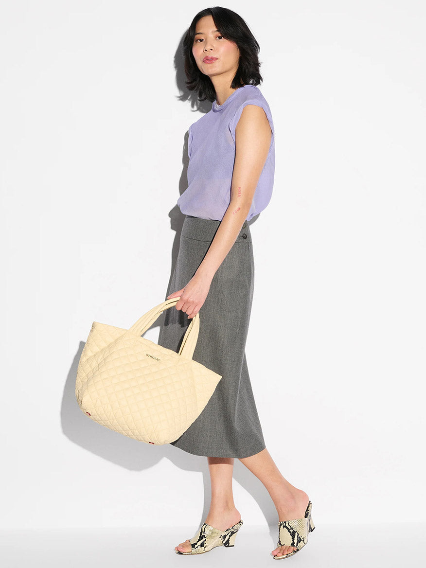 A woman in a lavender top, gray midi skirt, and patterned heeled sandals carries the MZ Wallace Small Metro Tote Deluxe in Chamomile Oxford against a plain white background.