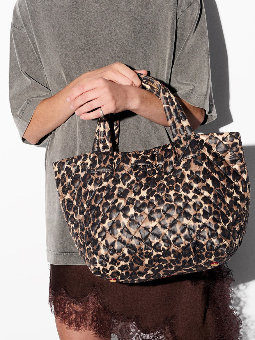 Person holding the MZ Wallace Small Metro Tote Deluxe in Leopard Oxford, featuring short handles and a crossbody strap, styled with a grey oversized t-shirt and a brown lace-trimmed skirt.