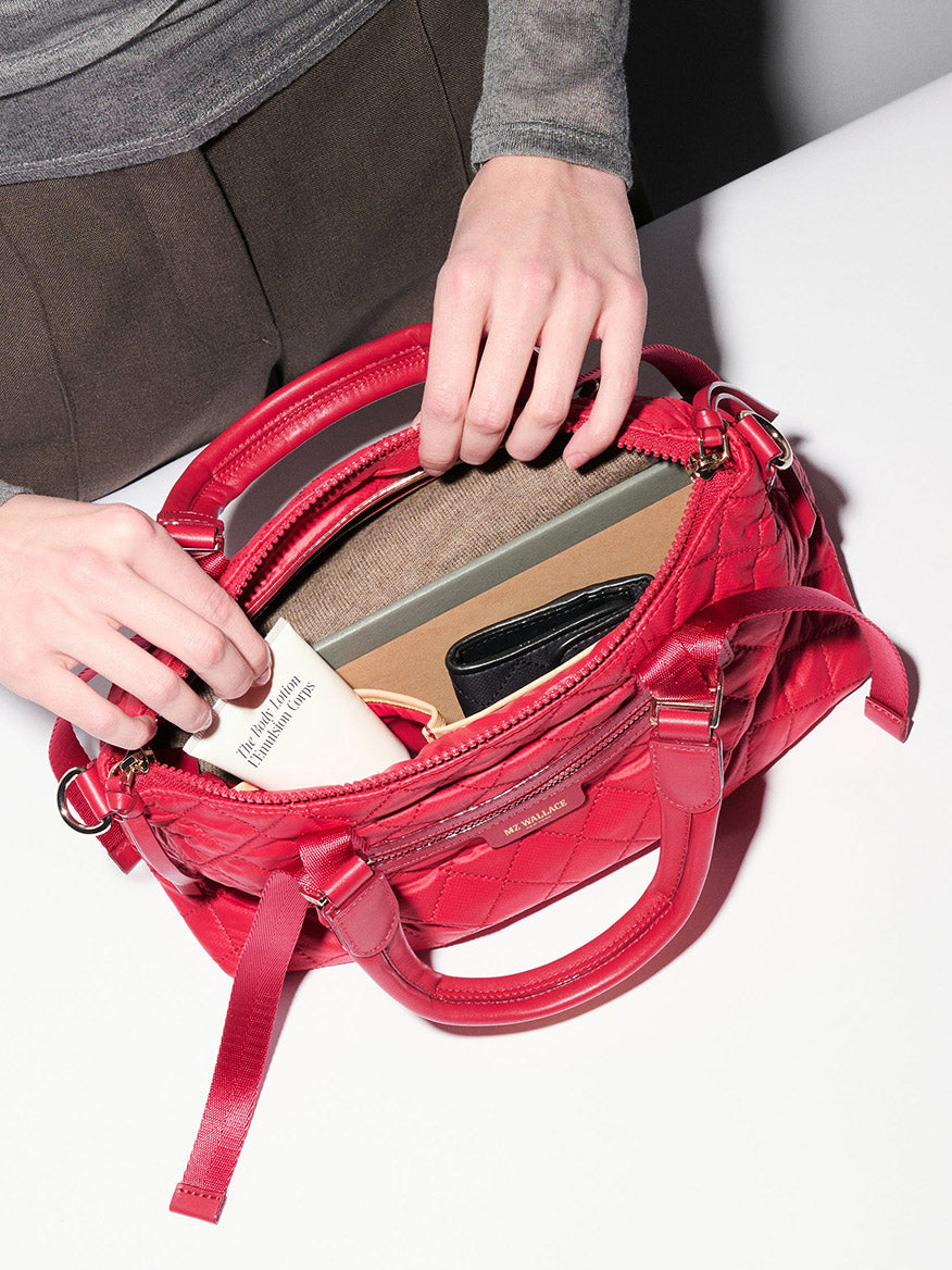 A person opens the MZ Wallace Small Park Satchel in Apple Oxford on a white surface, revealing a wallet, a book, and other personal items inside.