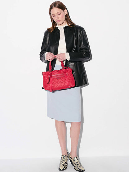 A woman stands against a white background wearing a black leather jacket, white blouse, light blue skirt, patterned ankle boots, and holding an MZ Wallace Small Park Satchel in Apple Oxford.