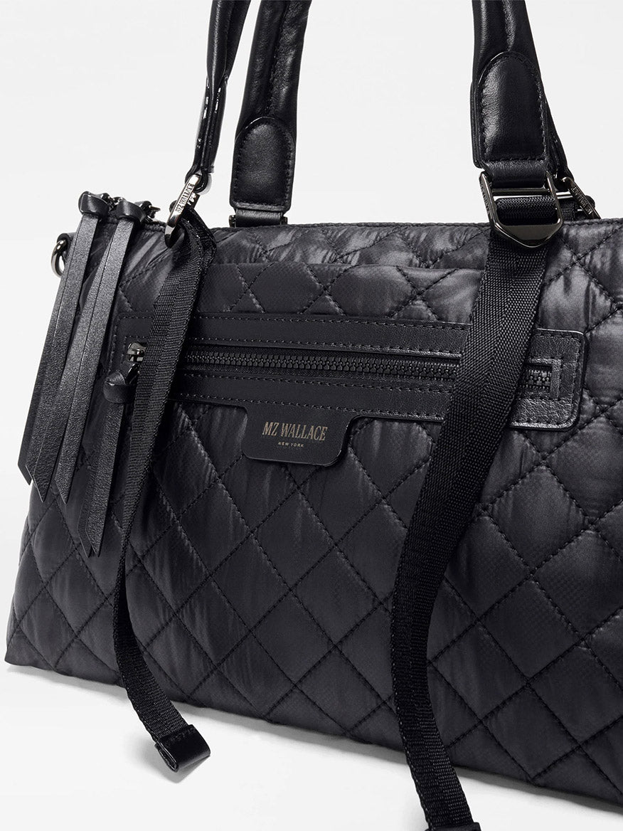 The MZ Wallace Small Park Satchel in Black Oxford boasts leather handles, a front zip pocket, and decorative tassels—combining stylish quilting and practicality for everyday use.