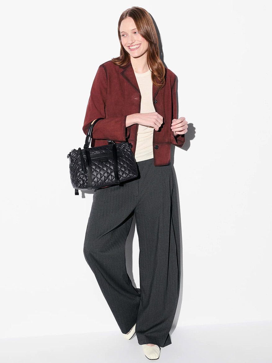 A woman stands against a white background wearing a maroon jacket, cream top, gray wide-leg trousers, white shoes, and holding the MZ Wallace Small Park Satchel in Black Oxford.