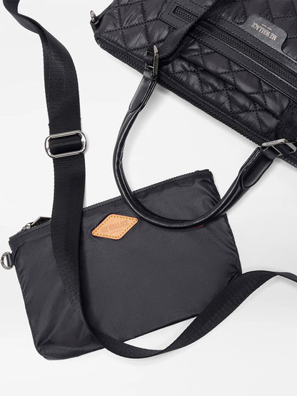 Two black bags are on a white surface: one is quilted with short handles; the other is the MZ Wallace Small Park Satchel in Black Oxford, featuring a plain design, brown patch, and long adjustable strap.