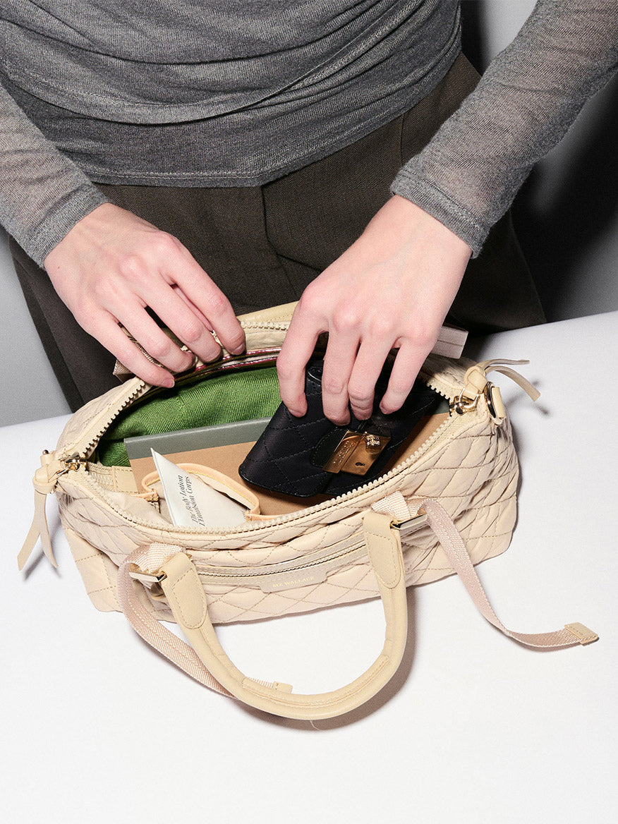 A person places a black wallet into an MZ Wallace Small Park Satchel in Buff Oxford, which features Italian leather trim and contains a green pouch and other small items, on a white surface.