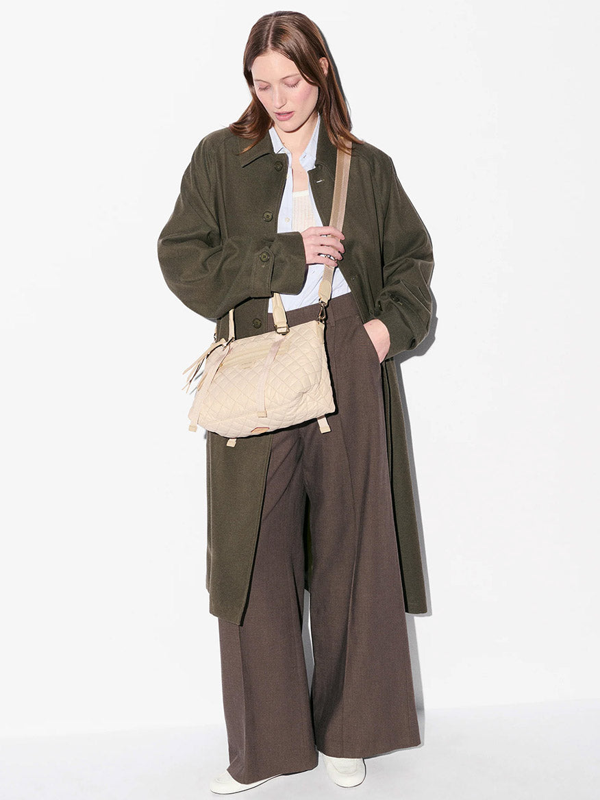 A person wearing a long dark green coat, wide brown pants, and white shoes stands against a white background, carrying the MZ Wallace Small Park Satchel in Buff Oxford.