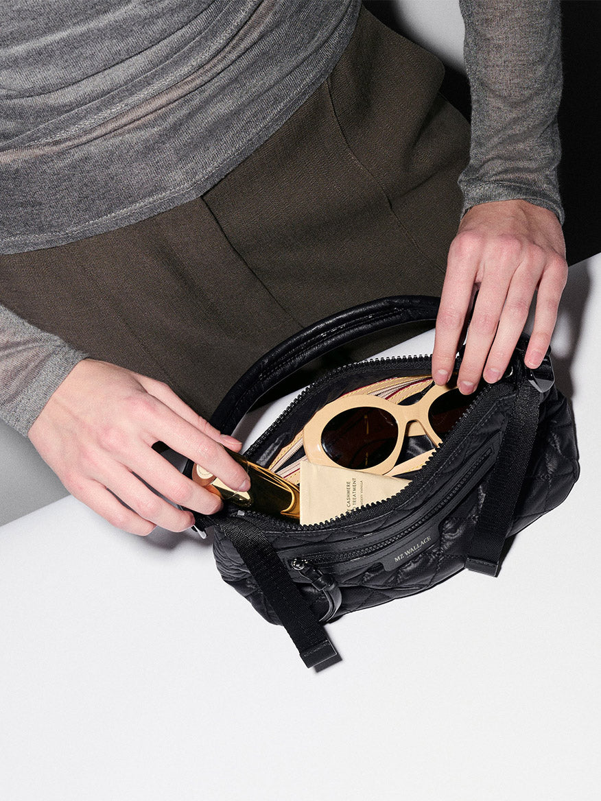 A person puts a pair of large round sunglasses and a gold lipstick tube into an MZ Wallace Small Park Shoulder Bag in Black Oxford on a white surface.