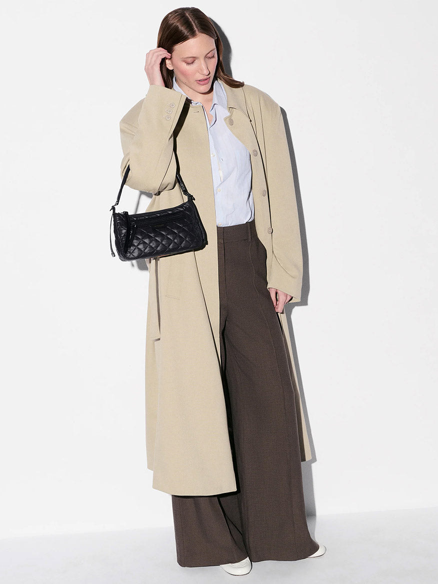 A woman stands against a white background wearing a light beige coat, light blue shirt, brown wide-leg pants, white shoes, and carrying the MZ Wallace Small Park Shoulder Bag in Black Oxford.