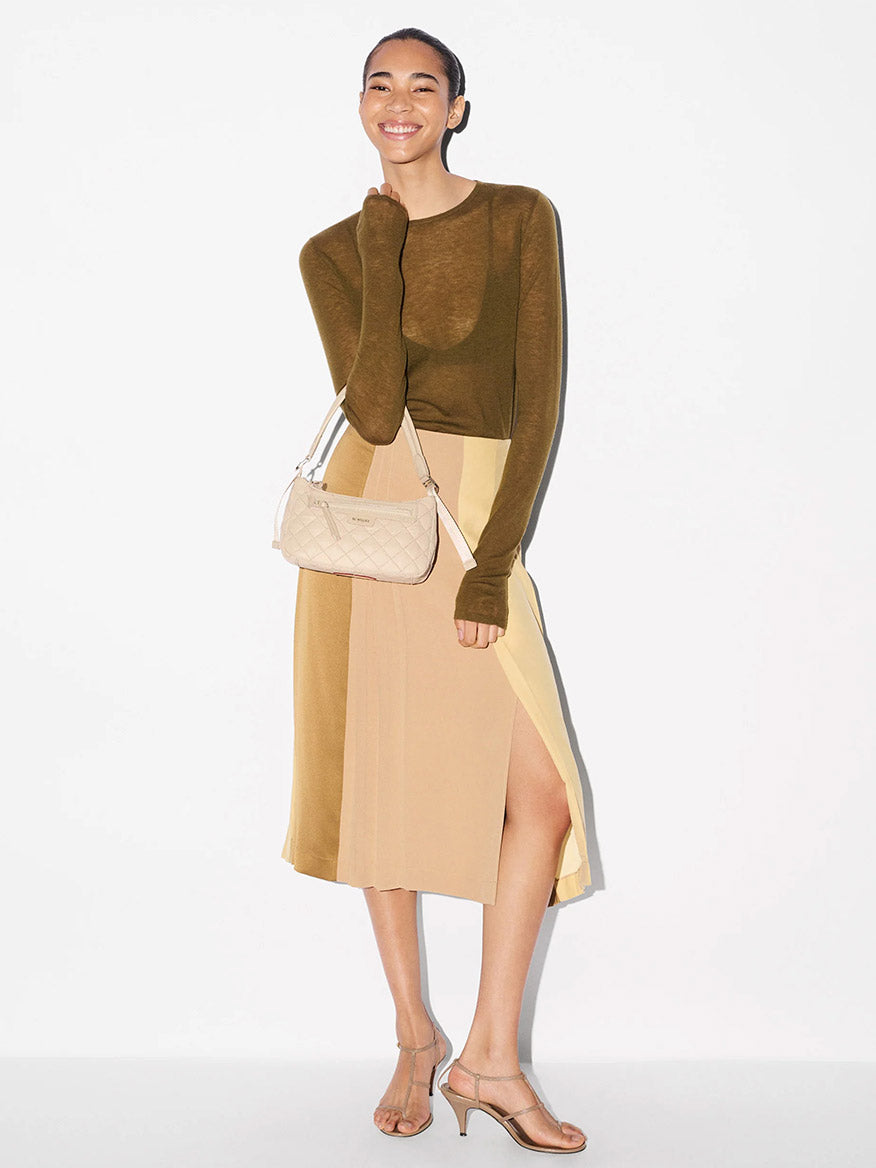 A woman stands smiling, wearing a long-sleeve olive top, beige colorblock skirt with a slit, strappy heeled sandals, and holding the MZ Wallace Small Park Shoulder Bag in Buff Oxford.