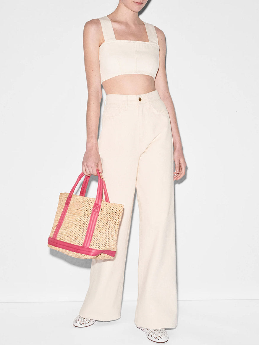 A woman in a stylish beige crop top and high-waisted trousers holding a MZ Wallace Small Raffia Tote in Raffia/Zinnia with padded nylon handles, against a plain white background.