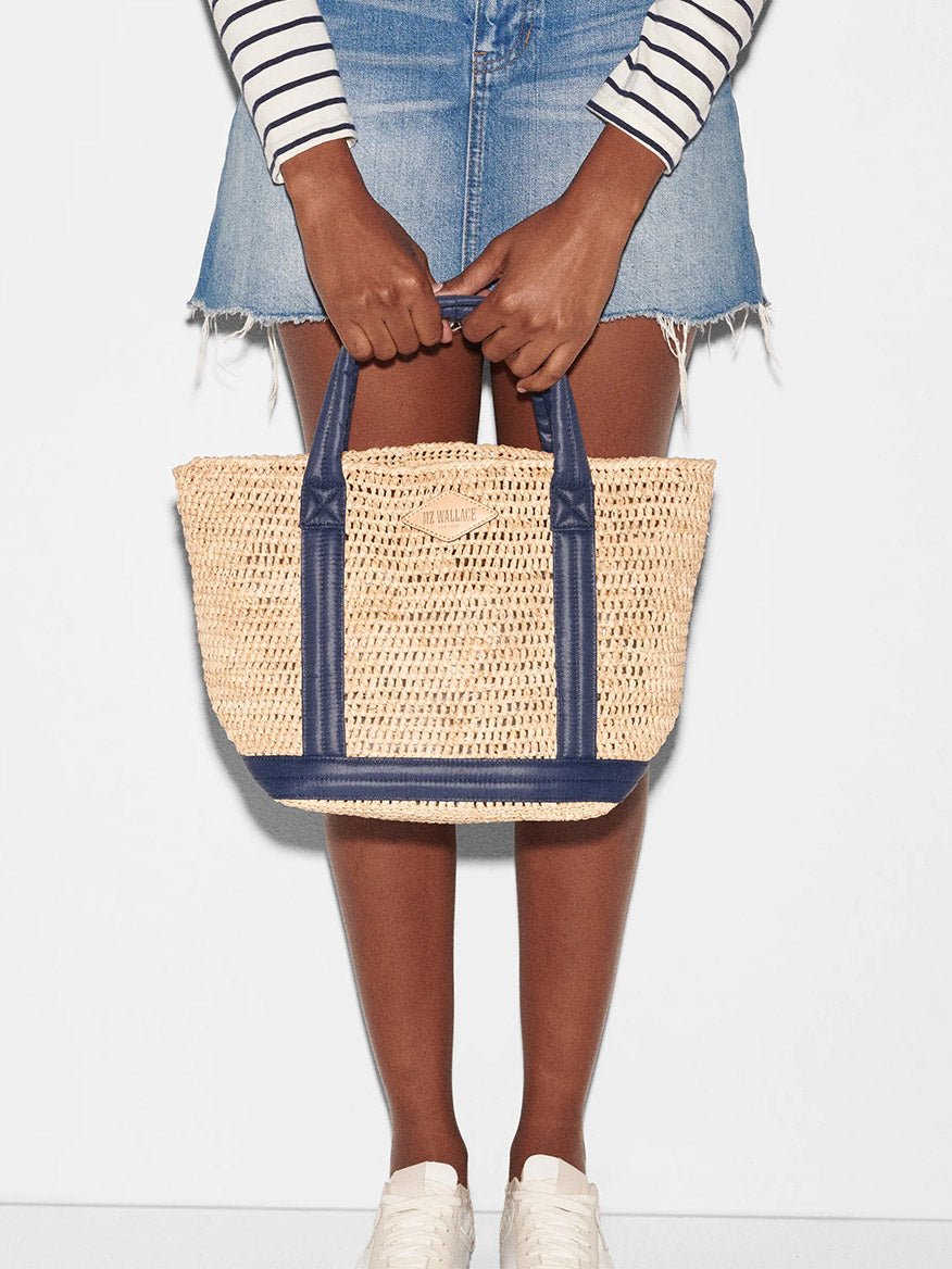 Person holding a MZ Wallace Small Raffia Tote in Raffia/Navy with padded nylon handles while standing against a white background.