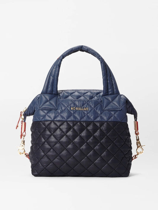 The MZ Wallace Small Sutton Deluxe in Black/Navy Color Block Oxford is a two-tone quilted handbag that boasts innovative features like short handles and a detachable crossbody strap, with a dark blue base, lighter blue top section, and gold-tone hardware.