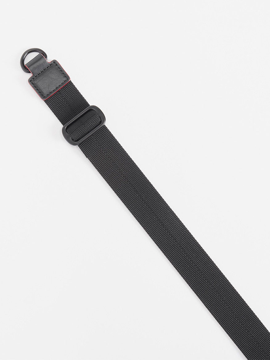 A black adjustable crossbody strap with a plastic buckle and metal D-ring is laid diagonally on a white surface as an accessory for the MZ Wallace Small Sutton Deluxe in Black Lacquer Oxford.