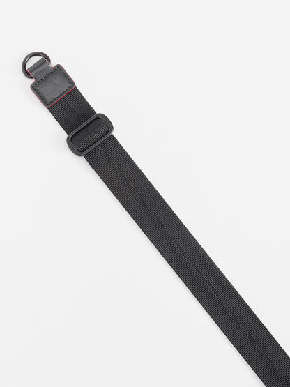 A black adjustable crossbody strap with a plastic buckle and metal D-ring is laid diagonally on a white surface as an accessory for the MZ Wallace Small Sutton Deluxe in Black Lacquer Oxford.