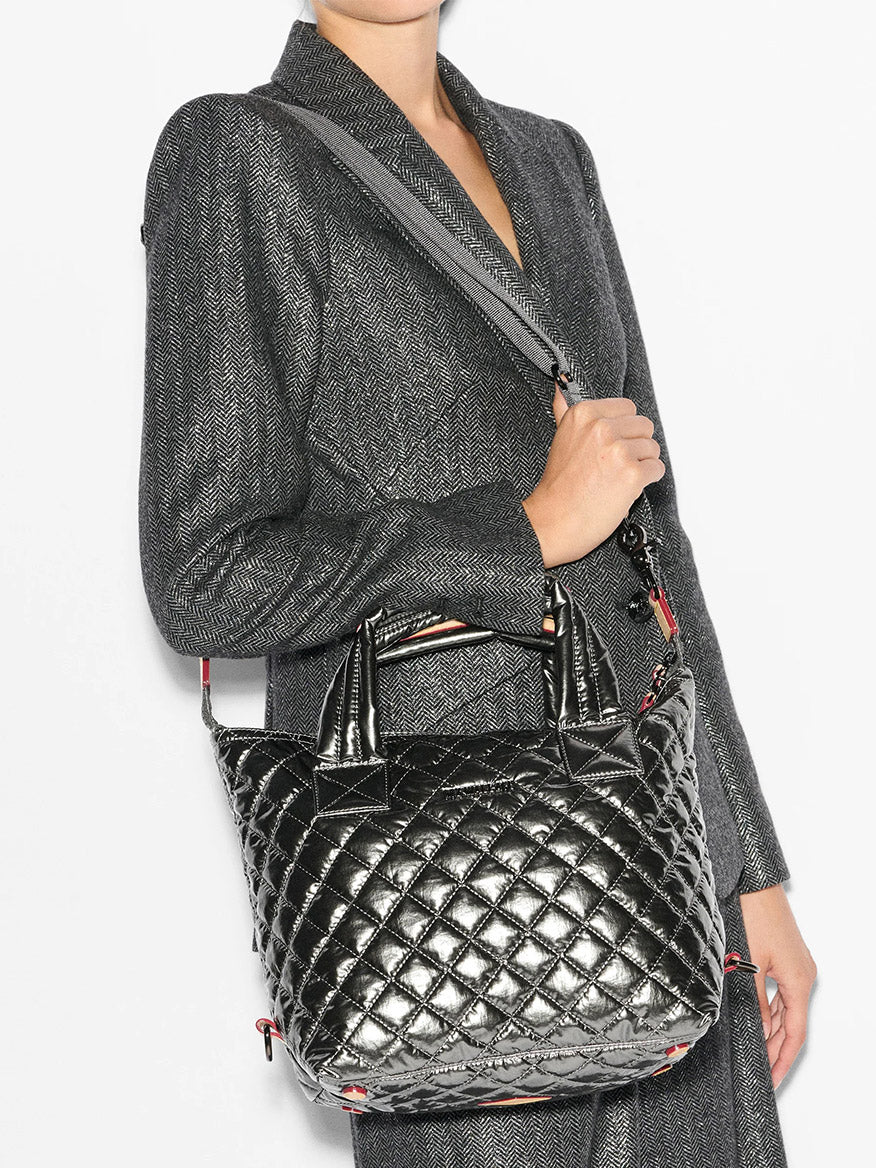 A person wearing a grey textured jacket holds the MZ Wallace Small Sutton Deluxe in Pewter Metallic Oxford, a shiny quilted handbag crafted from natural Italian leather, featuring a top handle and crossbody strap.