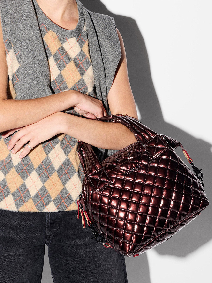 A person wearing a sleeveless argyle sweater vest and dark jeans holds the MZ Wallace Small Sutton Deluxe in Bordeaux Lacquer Oxford against a plain background.