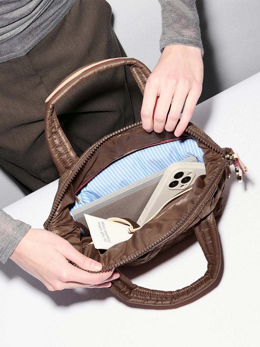 A person opens an MZ Wallace Small Sutton Deluxe in Walnut Oxford on a table, revealing a smartphone, a book, and a pair of glasses nestled inside its blue-striped interior crafted from recycled Oxford nylon.