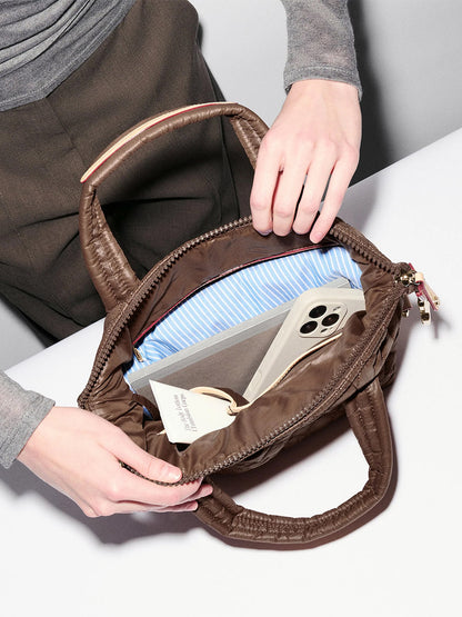 A person opens an MZ Wallace Small Sutton Deluxe in Walnut Oxford on a table, revealing a smartphone, a book, and a pair of glasses nestled inside its blue-striped interior crafted from recycled Oxford nylon.