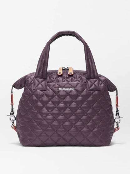 The MZ Wallace Small Sutton Deluxe in Raisin Oxford is a quilted dark purple handbag made from recycled Oxford nylon, featuring two short handles, a zip closure, and detachable shoulder strap hooks on each side, displayed against a plain white background.
