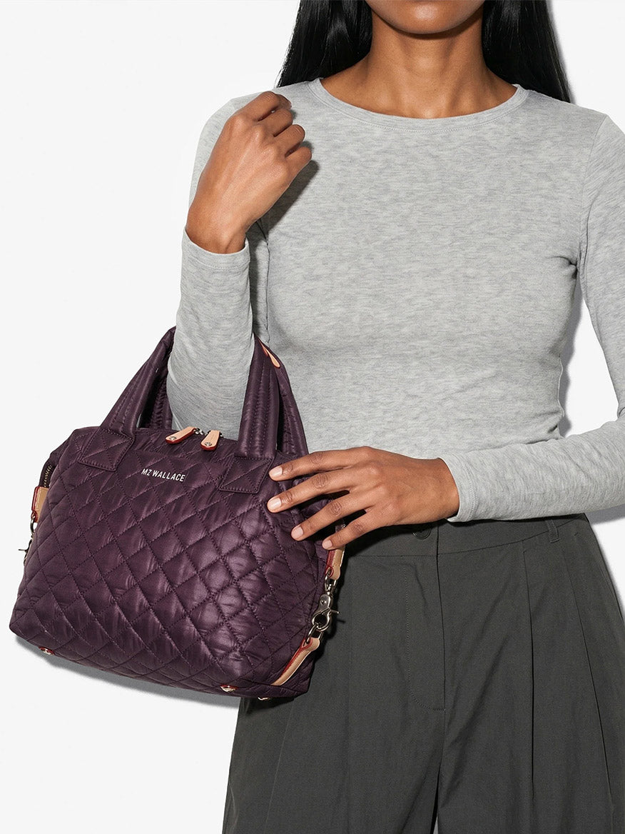 A person in a gray long-sleeve shirt and dark pants holds the MZ Wallace Small Sutton Deluxe in Raisin Oxford, a purple quilted handbag made from recycled Oxford nylon, featuring short handles, a detachable strap, and versatile compact crossbody styling.