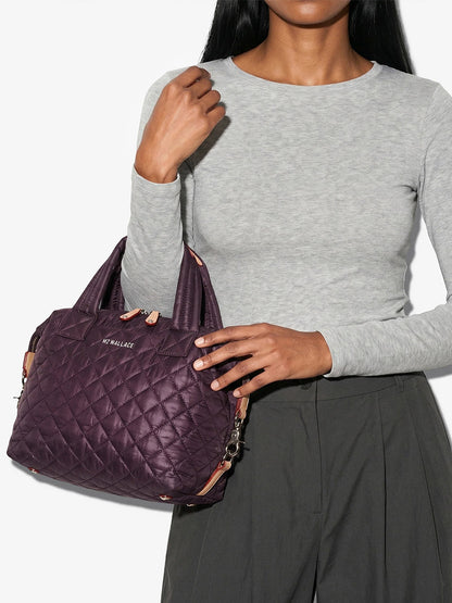 A person in a gray long-sleeve shirt and dark pants holds the MZ Wallace Small Sutton Deluxe in Raisin Oxford, a purple quilted handbag made from recycled Oxford nylon, featuring short handles, a detachable strap, and versatile compact crossbody styling.