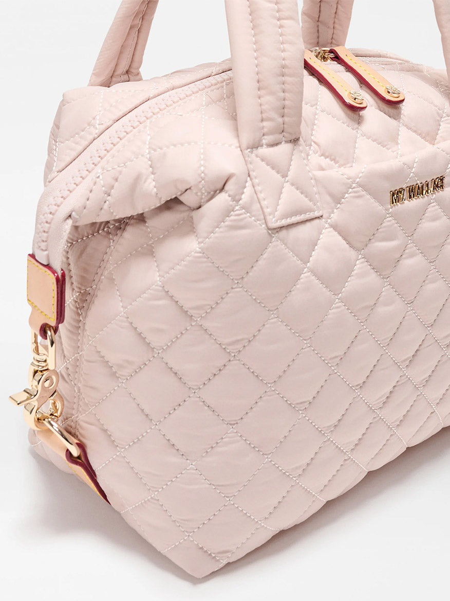 The MZ Wallace Small Sutton Deluxe in City Pink Oxford features quilted light pink material, dual handles, gold hardware, zip closure, adjustable crossbody strap, and a metal brand logo on the side.