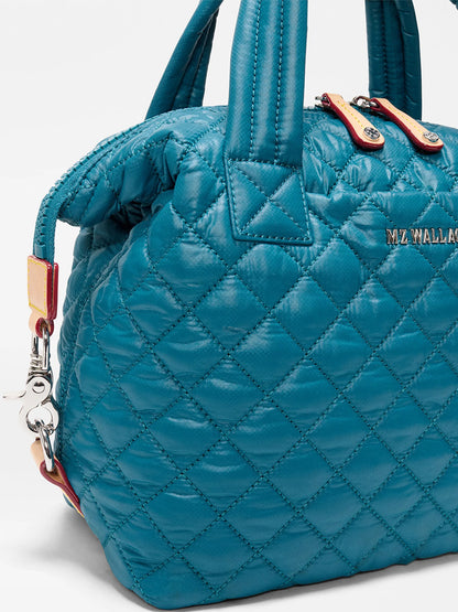 Close-up of the MZ Wallace Small Sutton Deluxe in Pacific Oxford, a blue quilted nylon bag with padded handles, an adjustable crossbody strap, metal zipper, and "MZ WALLACE" logo on the side.
