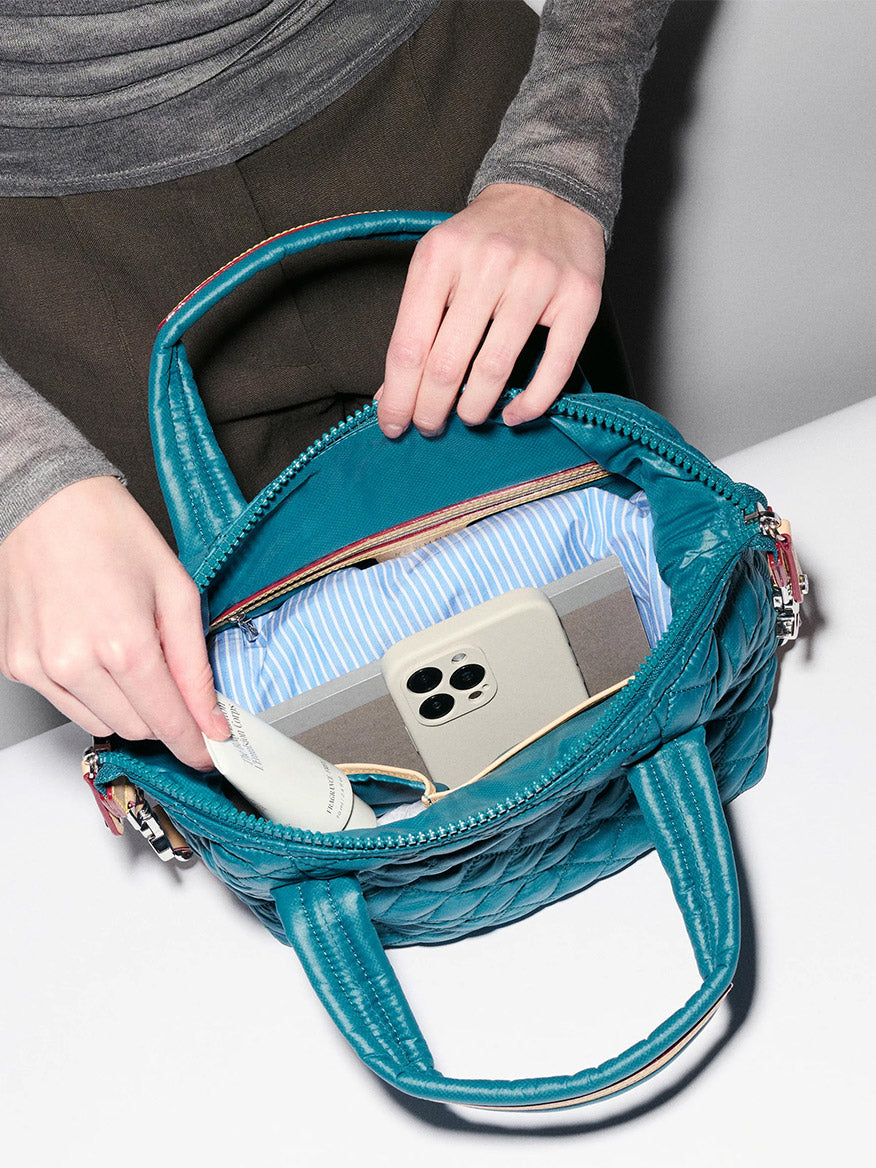 A person opens the MZ Wallace Small Sutton Deluxe in Pacific Oxford, revealing a smartphone, a striped pouch, and a white object inside; their hands and the bag’s adjustable crossbody strap are visible.