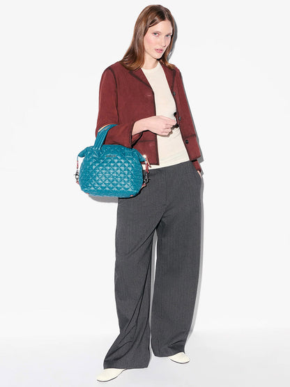 A person stands against a white background wearing a maroon jacket, cream top, gray wide-leg pants, white shoes, and carries the MZ Wallace Small Sutton Deluxe in Pacific Oxford quilted nylon bag with an adjustable crossbody strap.