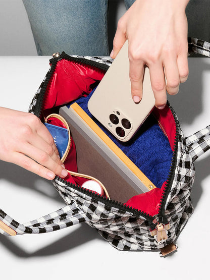 Someone puts a smartphone, a blue item, a brown notebook, and red-framed sunglasses into the MZ Wallace Small Sutton Deluxe in Black/White Gingham Oxford, which has an adjustable crossbody strap and Italian leather trim.