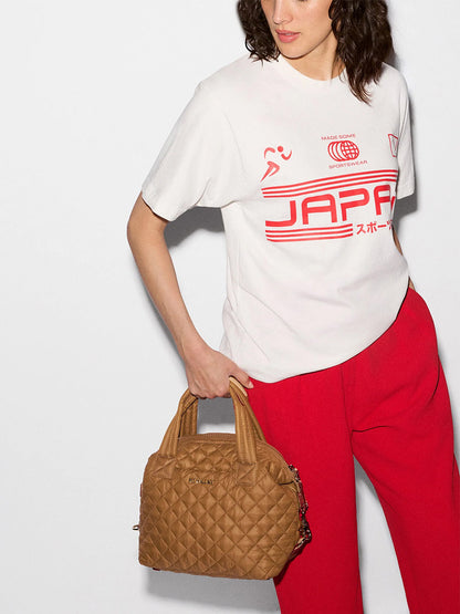 A person wearing a white graphic t-shirt and red trousers, carrying a small, quilted, tan-colored MZ Wallace Small Sutton Deluxe in Cognac Oxford with double handles.