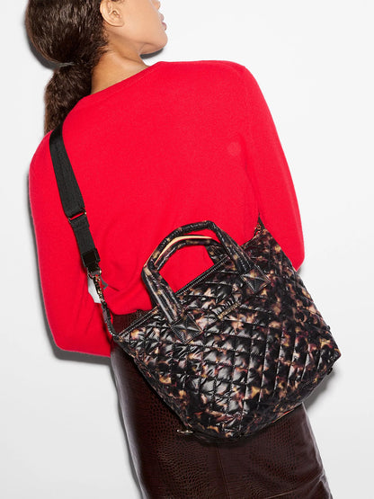 Person wearing a red sweater and brown skirt carrying an MZ Wallace Small Sutton Deluxe in Tortoise Shell Oxford, a small quilted crossbody tote with a brown and black tortoise shell print.