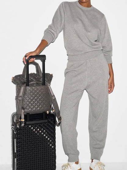 A person wearing a grey tracksuit stands holding the handle of a black patterned suitcase with a grey quilted bag on top. Their other hand rests on their hip. An MZ Wallace Small Utility Backpack in Magnet Oxford is slung over their shoulder.