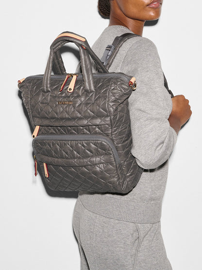 Person wearing a grey outfit and carrying an MZ Wallace Small Utility Backpack in Magnet Oxford, boasting 8 exterior pockets and a quilted brown design.