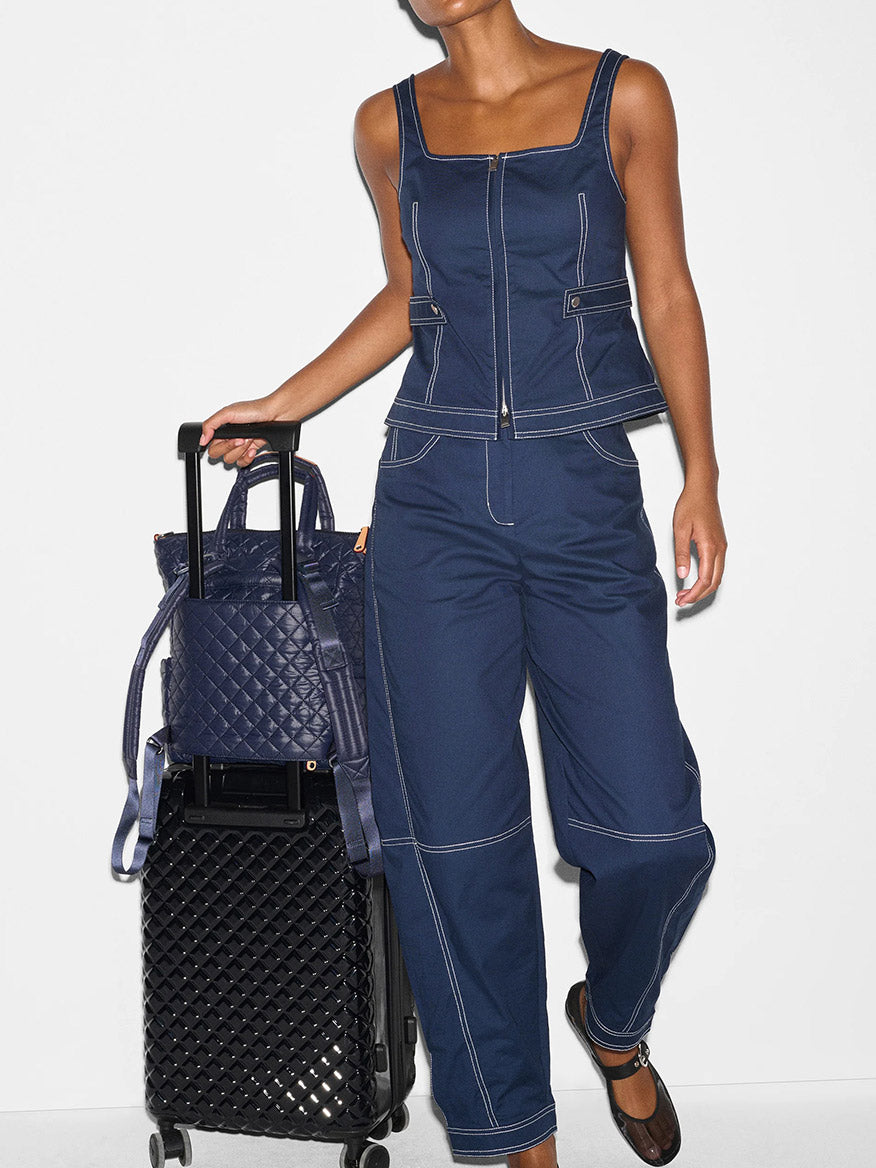 A person in a sleeveless denim jumpsuit stands against a white background, holding a quilted handbag and an MZ Wallace Small Utility Backpack in Dawn Oxford that fits a 14" laptop and is made from recycled oxford.