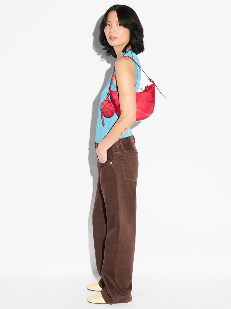 A person stands in profile against a white background wearing a light blue sleeveless top, brown wide-leg pants, beige shoes, and the MZ Wallace Small Waverly Hobo in Apple Bedford.