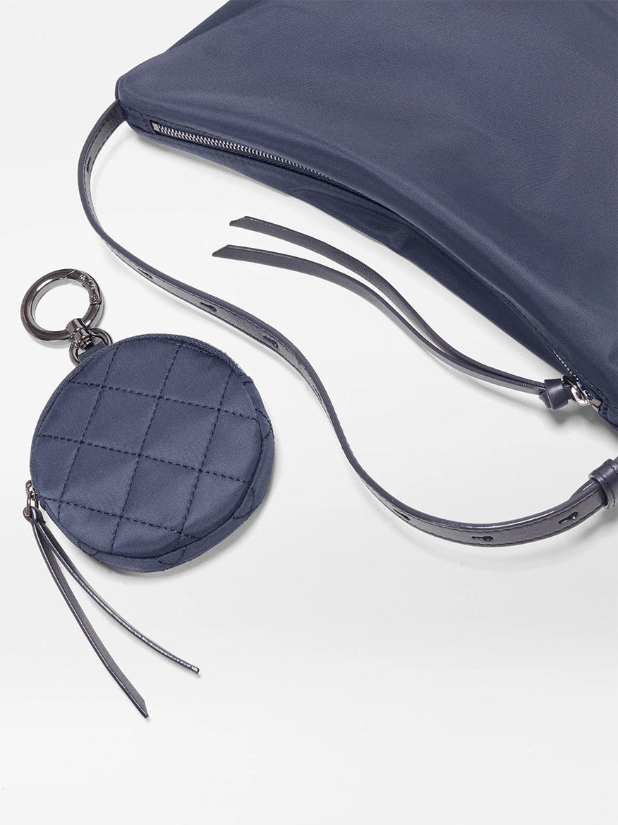 The MZ Wallace Small Waverly Hobo in Dawn Bedford, a navy blue bag with a long strap and round quilted coin pouch on a key ring, lies on a white surface.
